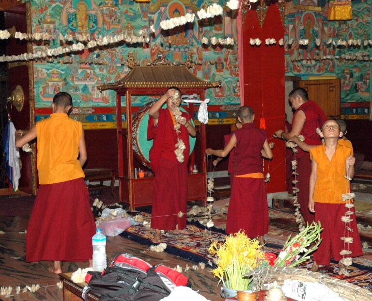 Young Buddhist monks in traditional robes engaged in decoration or ritual preparation inside a colorful monastery with Tibetan mural art on the walls.