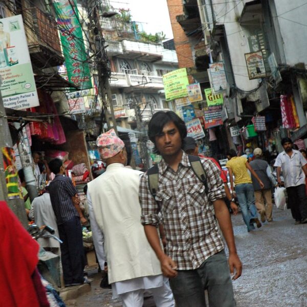 A bustling street scene in an urban market area with pedestrians, shops, and myriad signage. A young man with a shoulder bag walks in the foreground, looking to the side.