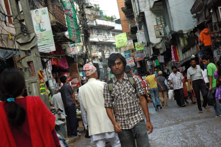 A bustling street scene in an urban market area with pedestrians, shops, and myriad signage. A young man with a shoulder bag walks in the foreground, looking to the side.