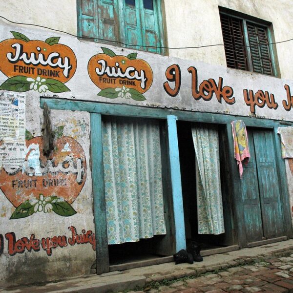 A faded building exterior with multiple "Juicy Fruit Drink" advertisements painted on its walls. There's a slogan saying "I love you Juicy" above a pair of wooden doors draped with worn curtains. Posters partially cover one advertisement, and laundry is hung to dry beside the doors.