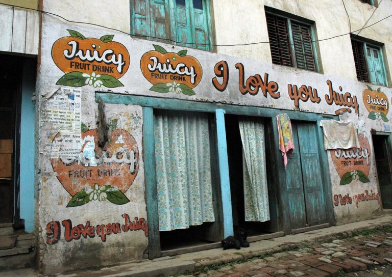 A faded building exterior with multiple "Juicy Fruit Drink" advertisements painted on its walls. There's a slogan saying "I love you Juicy" above a pair of wooden doors draped with worn curtains. Posters partially cover one advertisement, and laundry is hung to dry beside the doors.