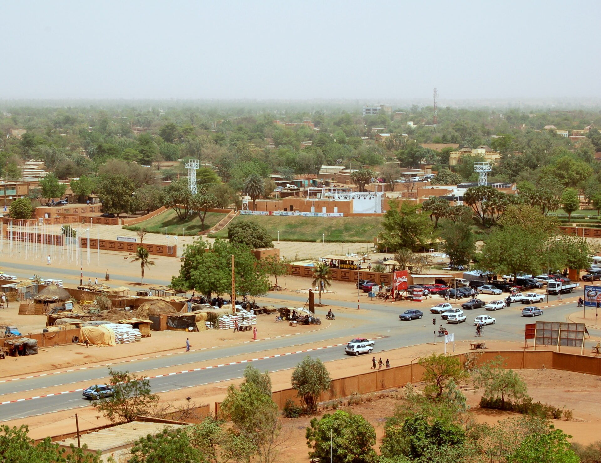 Aerial view of a bustling African town with dusty streets, a mix of traditional thatched stalls, modern buildings, and vehicles, under hazy skies.