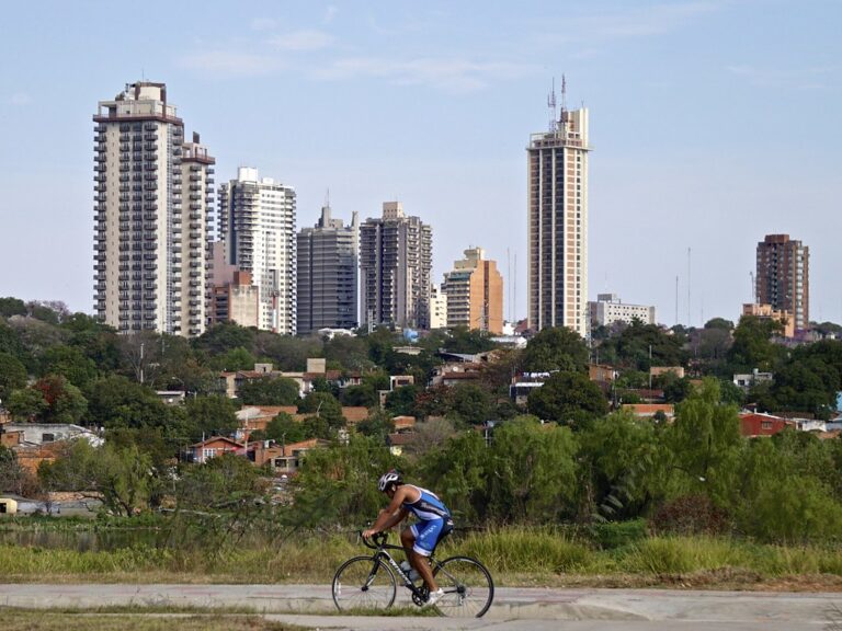 Cyclist in blue cycling gear riding on a path with a backdrop of various high-rise buildings and a clear sky.