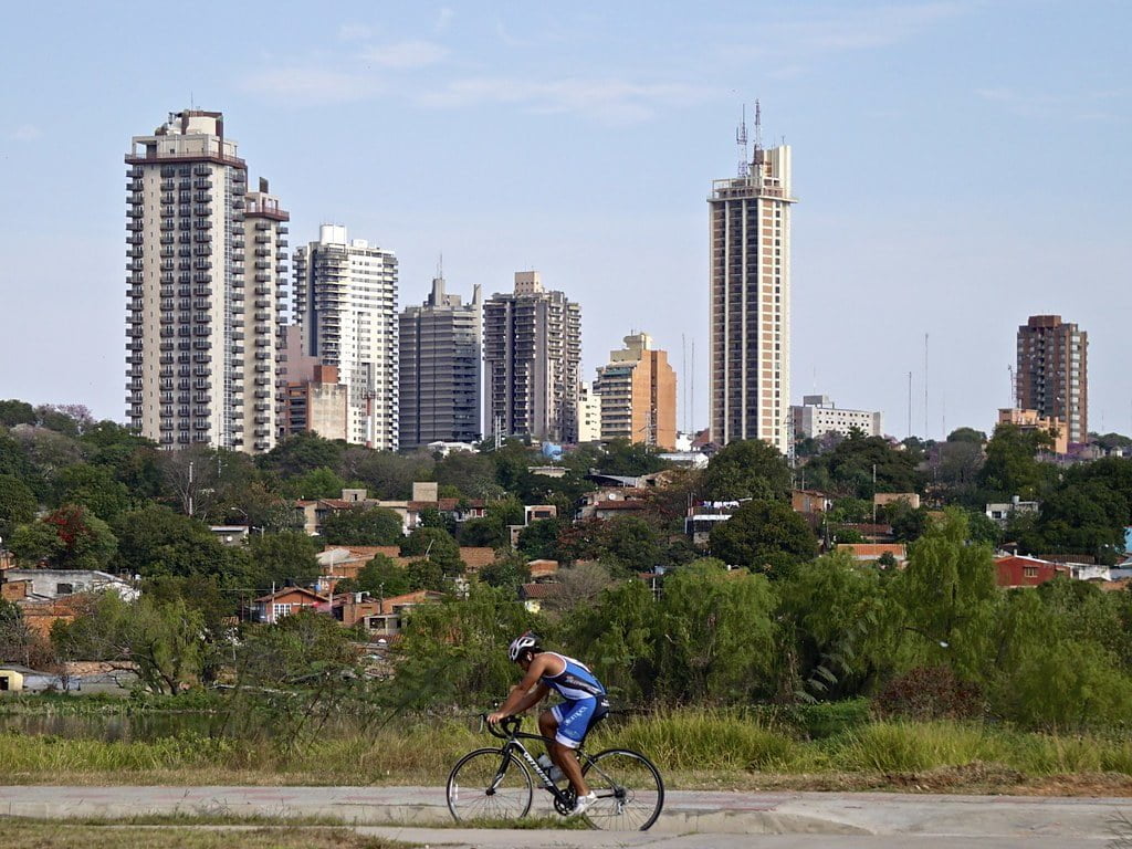 Cyclist in blue cycling gear riding on a path with a backdrop of various high-rise buildings and a clear sky.