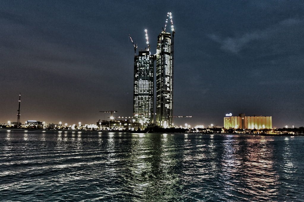 A nighttime cityscape featuring illuminated skyscrapers under construction beside calm water, with lights reflecting on the water's surface and other buildings in the background.