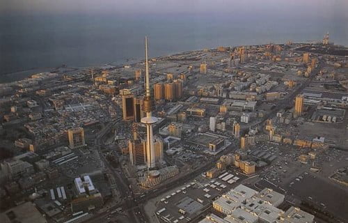 Aerial view of a cityscape at dusk with a prominent tower in the center and the coastline in the background.
