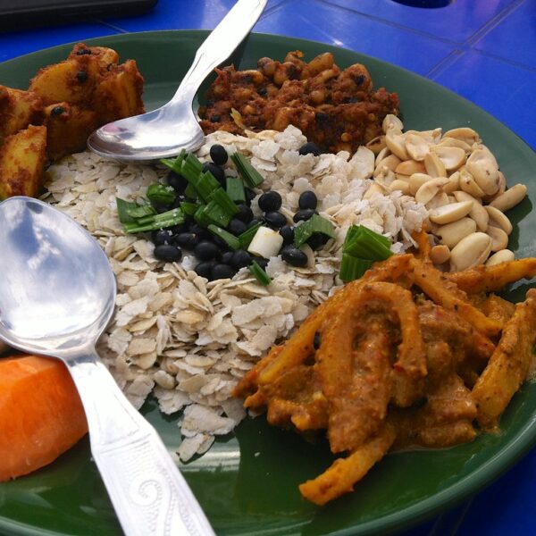 A green plate filled with various foods, including rice topped with spring onions and black beans, cucumber slices, carrots, fried potatoes, a spicy meat dish, and peanuts, served with a spoon on a blue table.