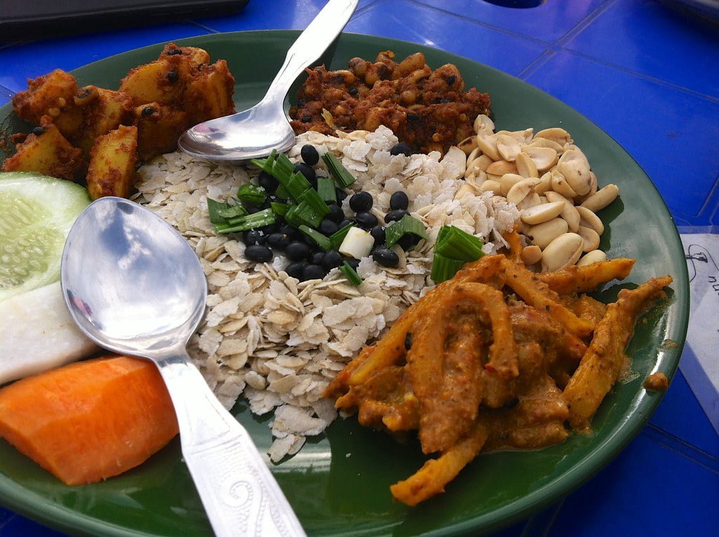 A green plate filled with various foods, including rice topped with spring onions and black beans, cucumber slices, carrots, fried potatoes, a spicy meat dish, and peanuts, served with a spoon on a blue table.