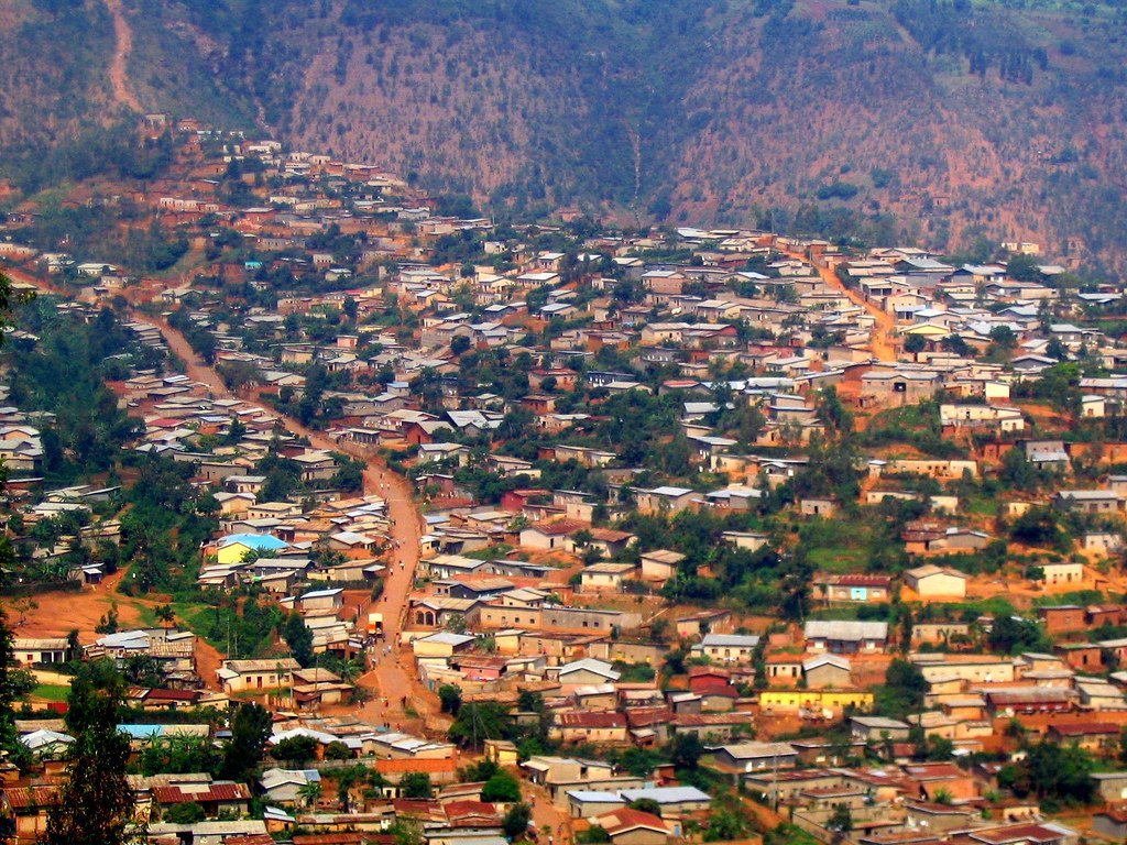 Aerial view of a densely populated town with reddish-brown rooftops, a winding dirt road down the center, nestled in the foothills of a mountainous area.