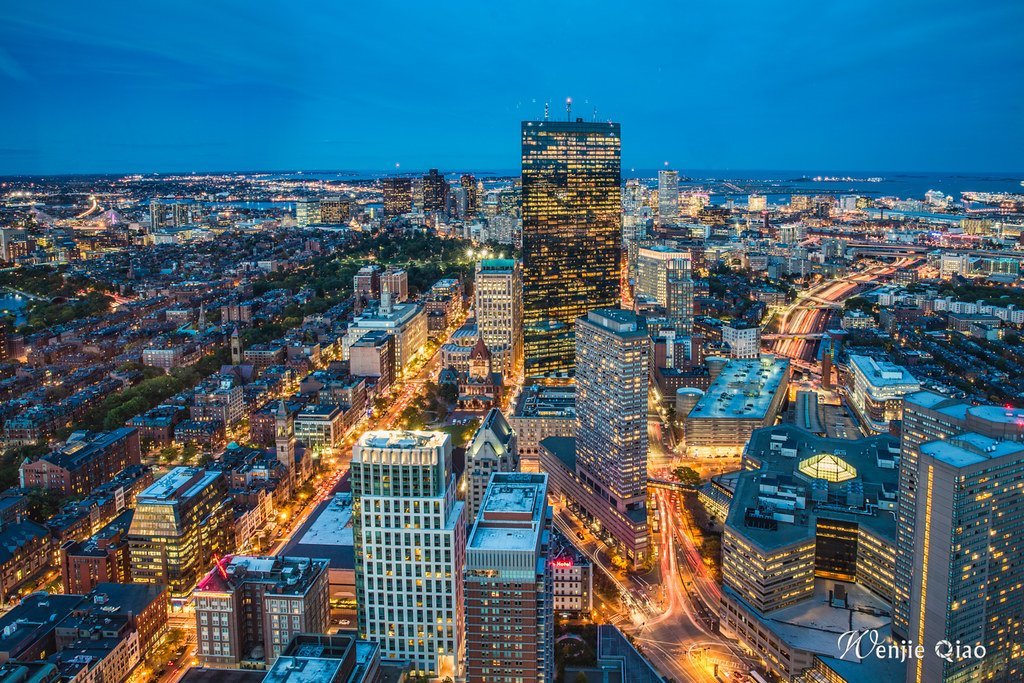 Alt text: Aerial view of a city at dusk with illuminated skyscrapers and busy streets, showcasing the transition from daylight to night with a vibrant blue sky and city lights.