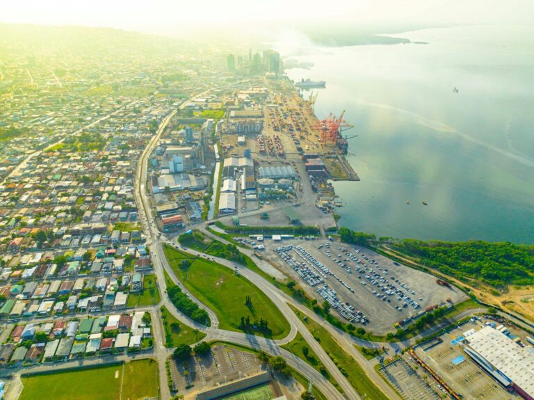 Aerial view of a coastal urban area with a port, shipping containers, roadways, and dense residential housing, with a body of water and hazy sky in the background.