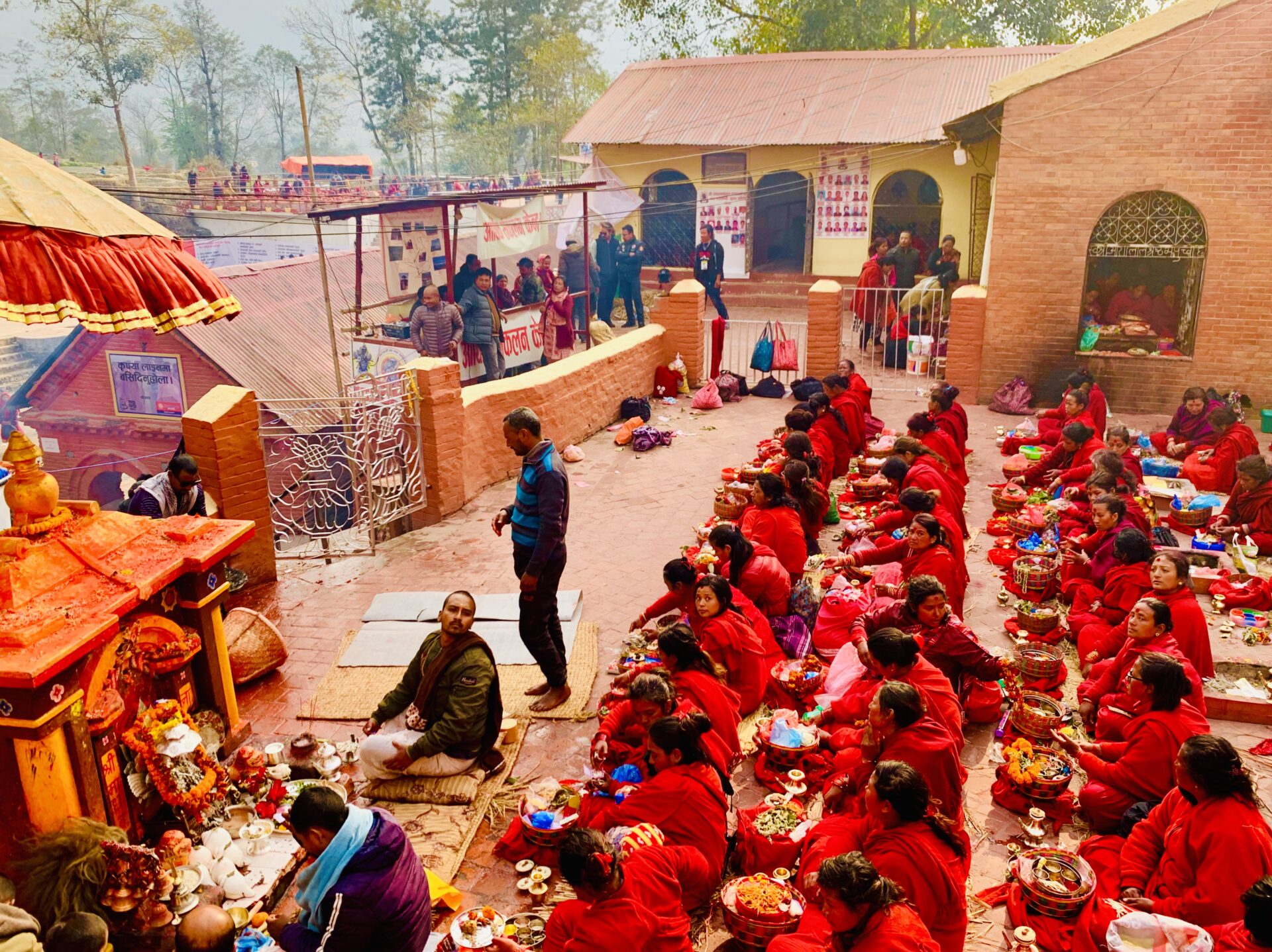A group of people in red attire sitting on the ground performing a religious ritual with various offerings in bowls at an outdoor temple complex, with onlookers standing at the fence.