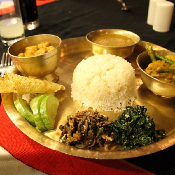 A traditional Indian meal served on a brass thali, including white rice, various curries, fried bread, slices of cucumber, and sautéed greens.
