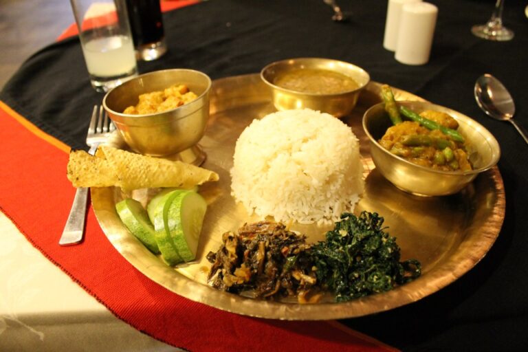 A traditional Indian meal served on a brass thali, including white rice, various curries, fried bread, slices of cucumber, and sautéed greens.