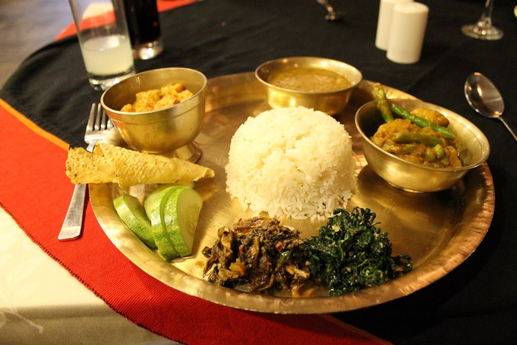 A traditional Indian meal served on a brass thali, including white rice, various curries, fried bread, slices of cucumber, and sautéed greens.