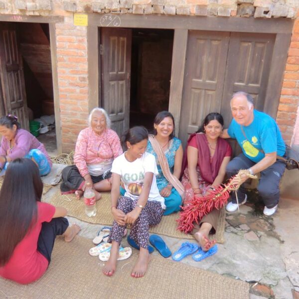 A group of people, both men and women, sit in front of a brick building with wooden doors, smiling and working with dried plants on mats. A man on the right kneels, holding a red plant.