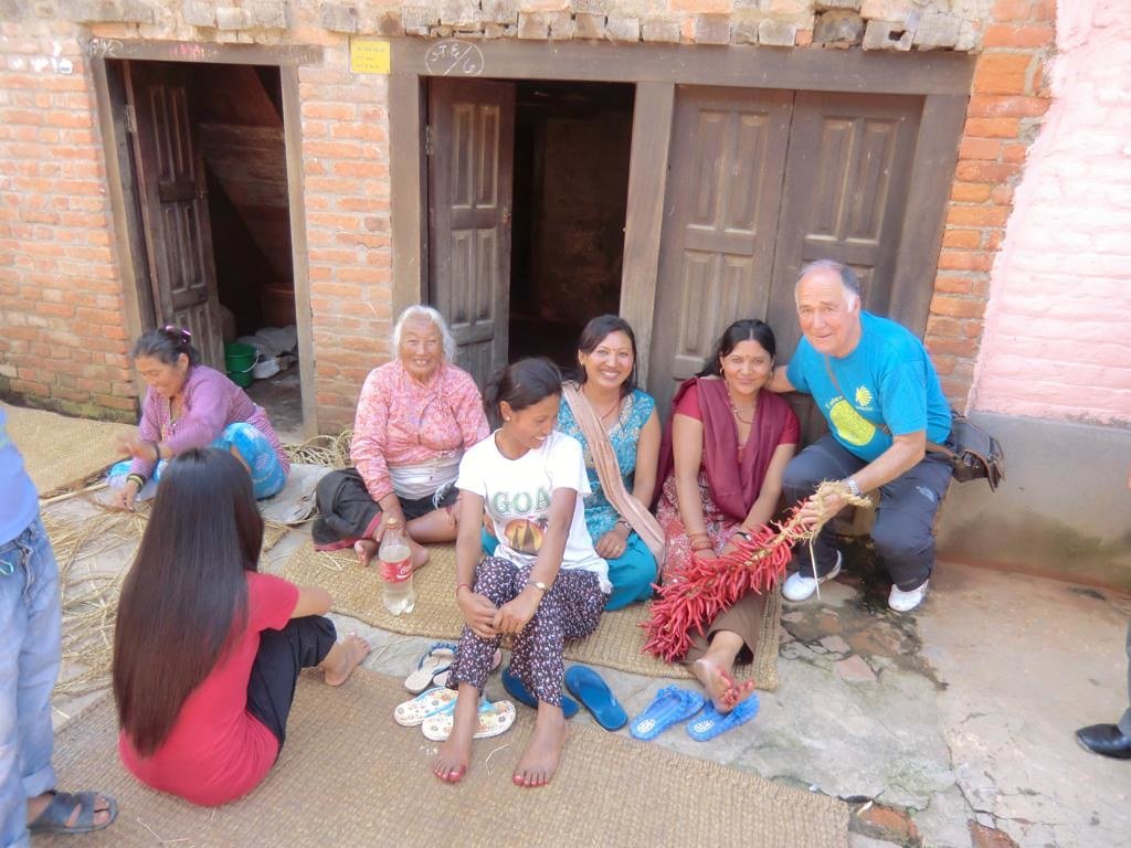 A group of people, both men and women, sit in front of a brick building with wooden doors, smiling and working with dried plants on mats. A man on the right kneels, holding a red plant.