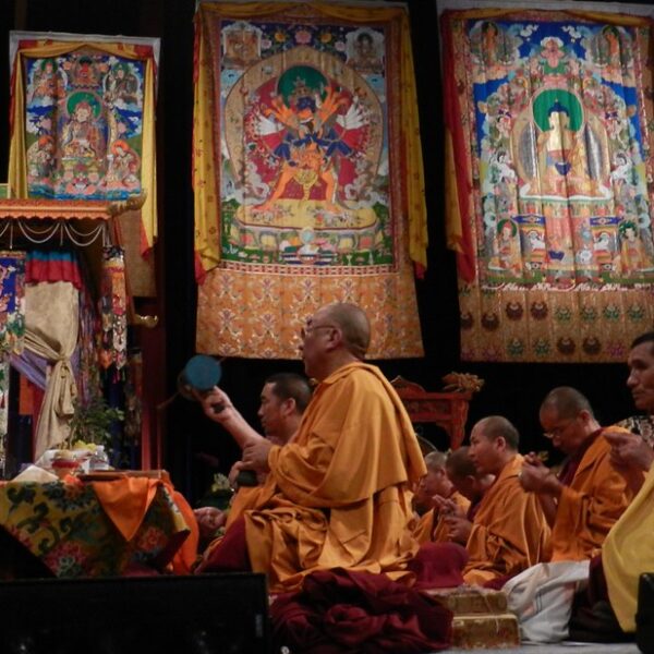 Monks in traditional yellow and red robes participating in a ceremony with colorful Tibetan thangka paintings hanging in the background.