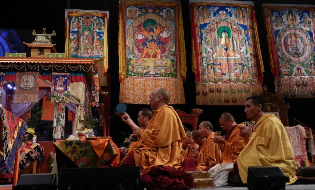 Monks in traditional yellow and red robes participating in a ceremony with colorful Tibetan thangka paintings hanging in the background.