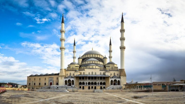 A large mosque with multiple domes and slender minarets against a partly cloudy sky, with an expansive paved courtyard in the foreground.