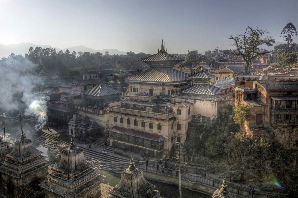 Aerial view of a historic temple complex with traditional architecture, showing smoke rising amidst the buildings, set against a backdrop of distant mountains.