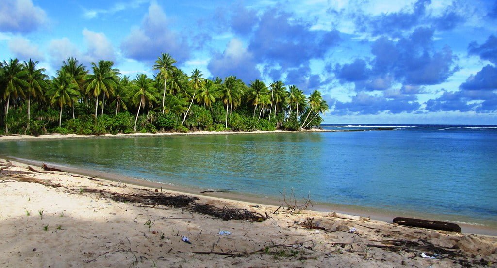 A serene tropical beach with clear blue waters, lined with lush green palm trees, under a partly cloudy sky.