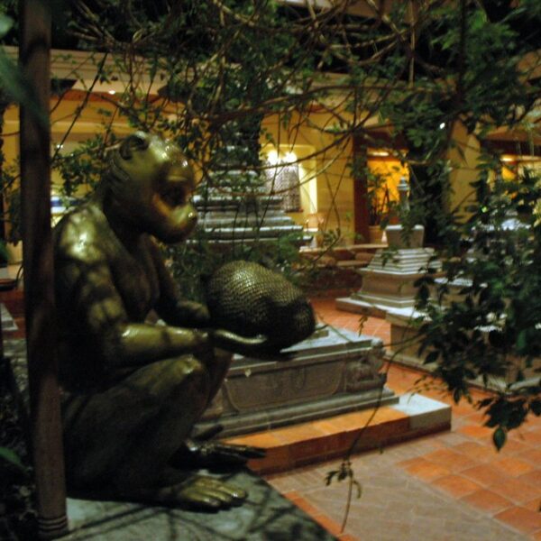 A bronze statue of a seated humanoid figure holding a spherical object, set in an indoor garden with terracotta tiles, surrounded by plants and background decorative elements that suggest a lobby or atrium area.