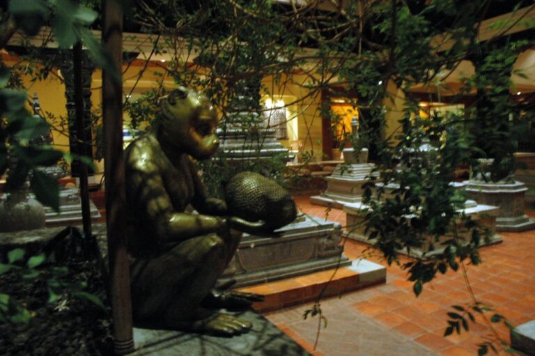 A bronze statue of a seated humanoid figure holding a spherical object, set in an indoor garden with terracotta tiles, surrounded by plants and background decorative elements that suggest a lobby or atrium area.
