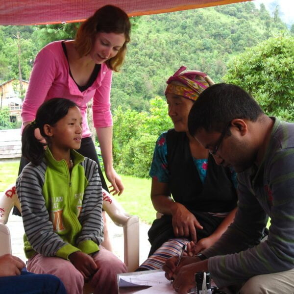 A group of people, including a young girl sitting and adults standing and sitting around a table, with a focus on a writing activity outdoors under a tent with lush greenery in the background.