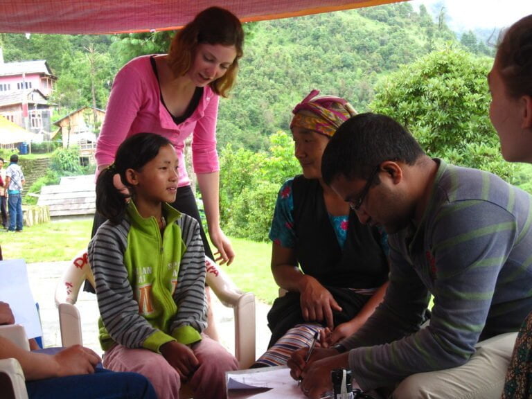 A group of people, including a young girl sitting and adults standing and sitting around a table, with a focus on a writing activity outdoors under a tent with lush greenery in the background.