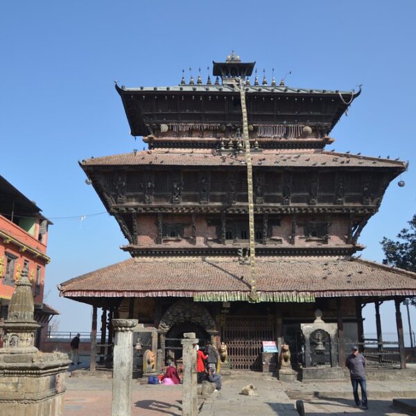 A multi-tiered pagoda-style temple with ornate wood carvings stands under a clear blue sky, surrounded by a few smaller structures and a couple of people around the temple's plaza.