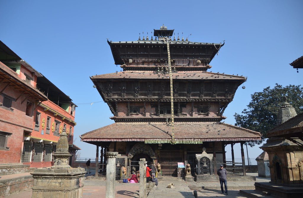 A multi-tiered pagoda-style temple with ornate wood carvings stands under a clear blue sky, surrounded by a few smaller structures and a couple of people around the temple's plaza.