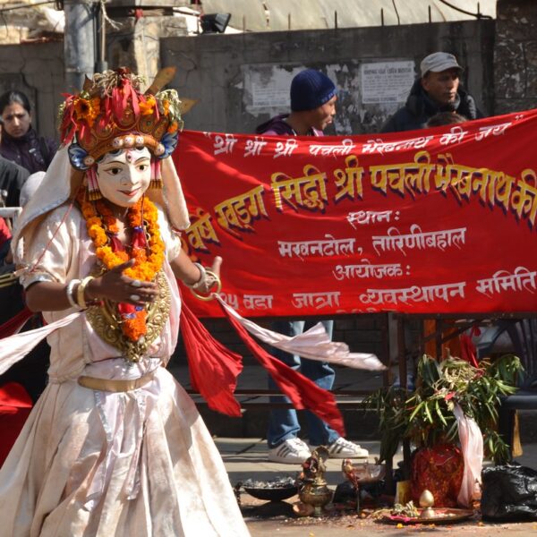 A performer dressed in traditional attire and elaborate facial make-up resembling a Hindu deity dances in front of a red banner with Devanagari script, during a cultural festival in India. Spectators watch from behind barricades.