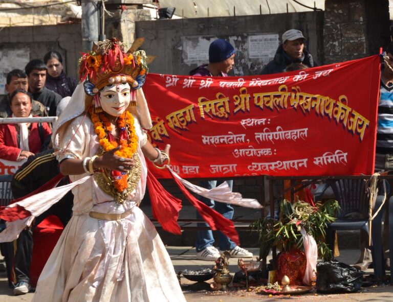 A performer dressed in traditional attire and elaborate facial make-up resembling a Hindu deity dances in front of a red banner with Devanagari script, during a cultural festival in India. Spectators watch from behind barricades.