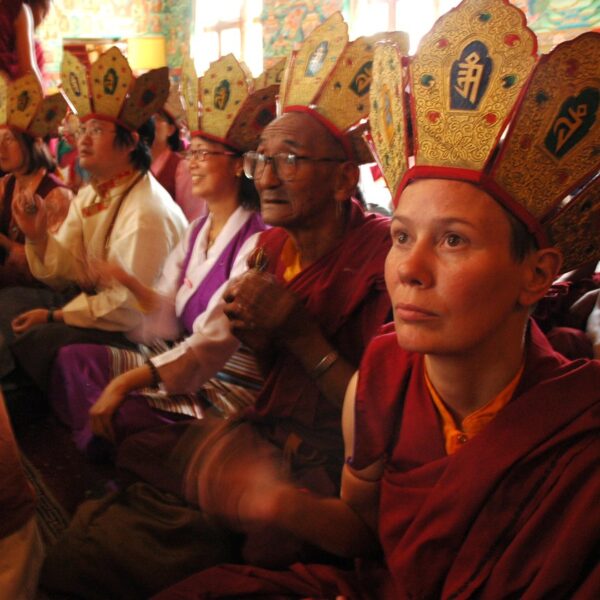 Group of people, including monks in traditional Tibetan Buddhist attire with elaborate hats, gathered in a room with colorful murals, participating in a ceremony.