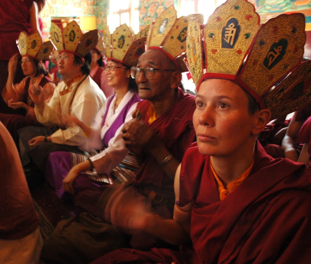 Group of people, including monks in traditional Tibetan Buddhist attire with elaborate hats, gathered in a room with colorful murals, participating in a ceremony.