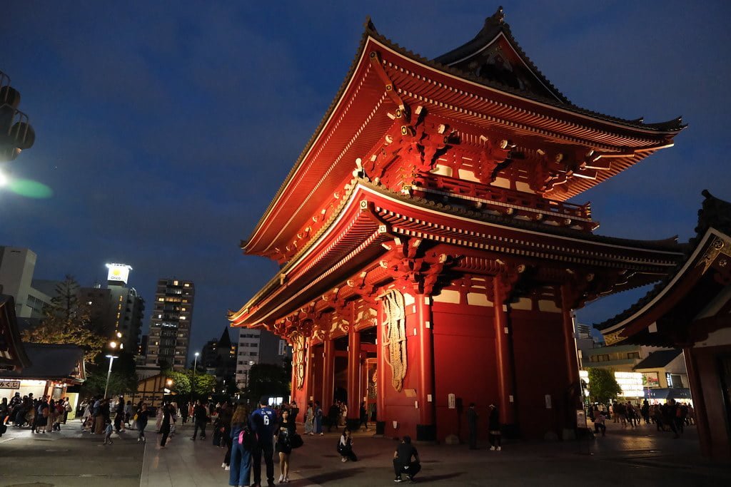 Alt text: A vibrant red traditional Japanese gate known as a torii, illuminated at twilight with people walking around the temple grounds. Skyscrapers rise in the background, signaling an urban setting.