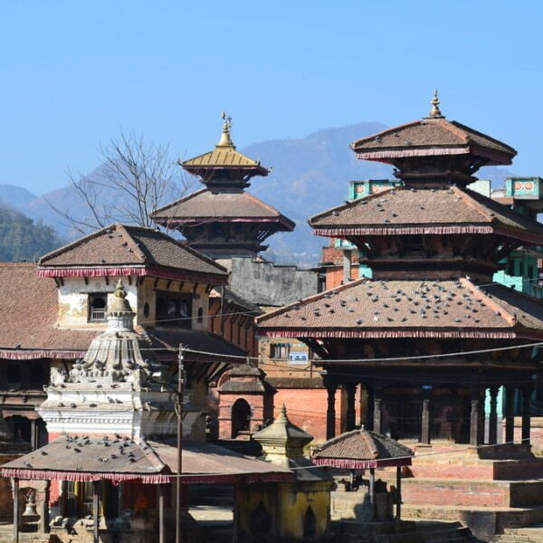 Traditional Nepalese pagoda-style temples and structures with tiered roofs in the foreground and mountainous landscape in the background under a clear blue sky.