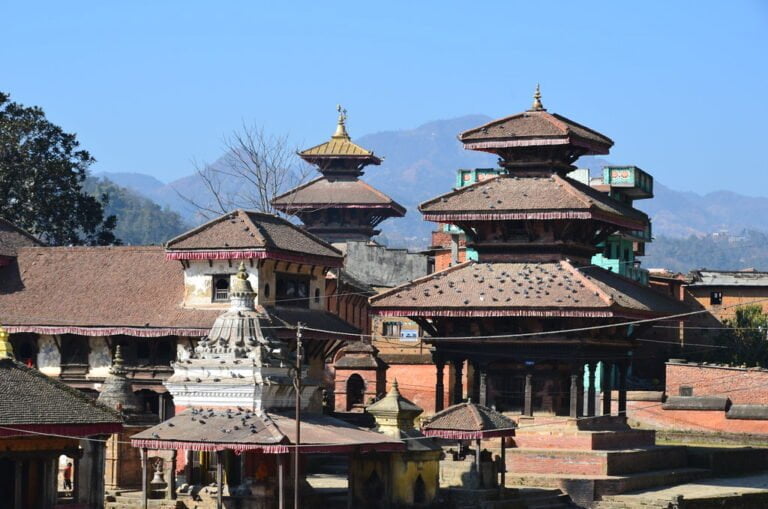 Traditional Nepalese pagoda-style temples and structures with tiered roofs in the foreground and mountainous landscape in the background under a clear blue sky.