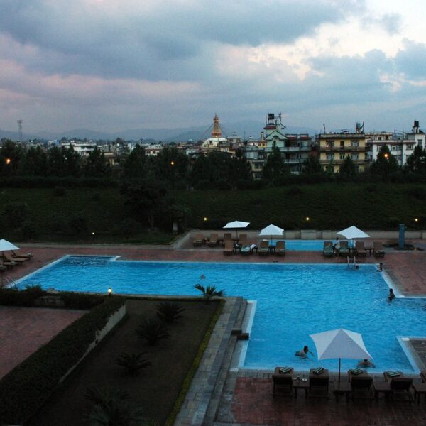 A twilight view of an outdoor swimming pool at a luxury hotel with lounge chairs and umbrellas, overlooked by a city with buildings against a cloudy sky.