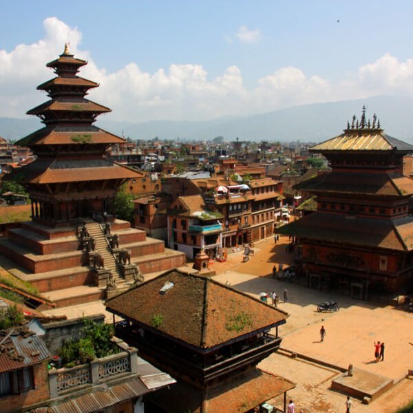 A panoramic view of two traditional Nepalese pagoda-style temples with tiered roofs in an expansive square surrounded by densely packed brick houses, set against a backdrop of distant hills and a clear sky.