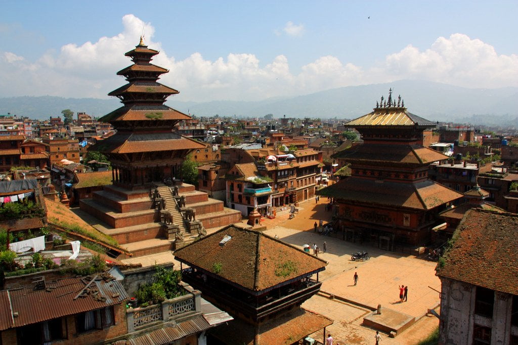 A panoramic view of two traditional Nepalese pagoda-style temples with tiered roofs in an expansive square surrounded by densely packed brick houses, set against a backdrop of distant hills and a clear sky.