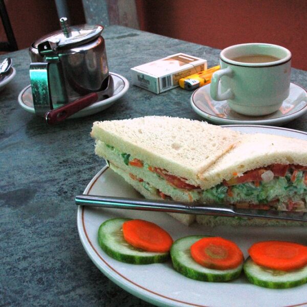 A table with a meal consisting of a sandwich on a plate with cucumber slices, a cup of tea, a tea set, and a pack of cigarettes.