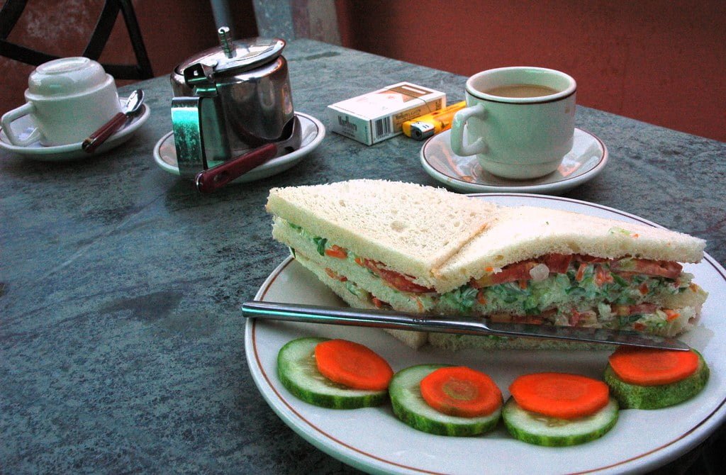 A table with a meal consisting of a sandwich on a plate with cucumber slices, a cup of tea, a tea set, and a pack of cigarettes.