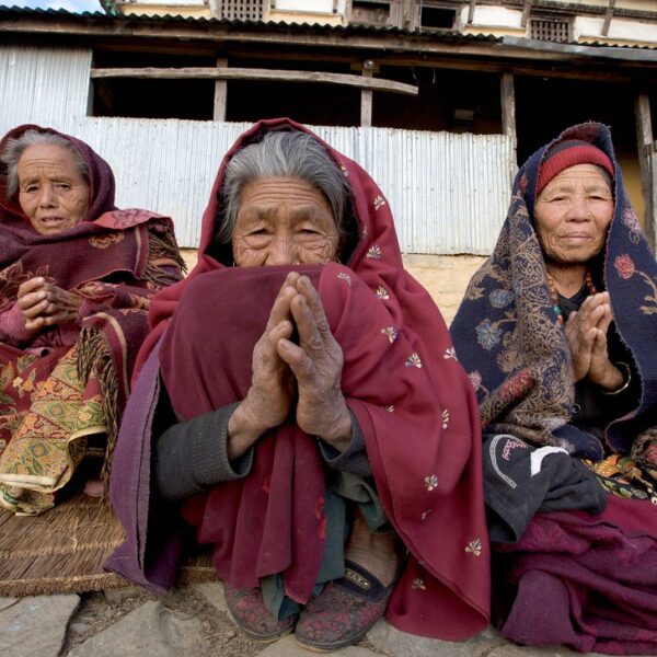 Three elderly women in traditional attire sitting together with their hands pressed together in a gesture of greeting or prayer, in front of a rustic building with metal sheeting.
