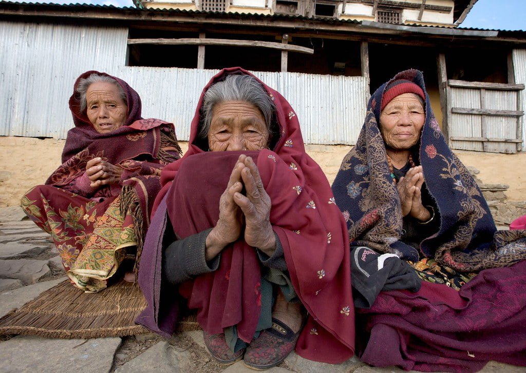 Three elderly women in traditional attire sitting together with their hands pressed together in a gesture of greeting or prayer, in front of a rustic building with metal sheeting.