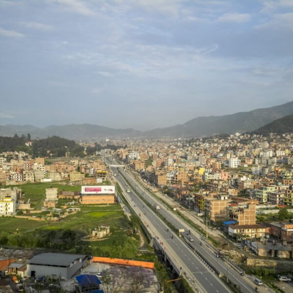 Aerial view of a bustling city with dense building clusters, a clear highway cutting through the middle, and surrounding green hills under a blue sky.
