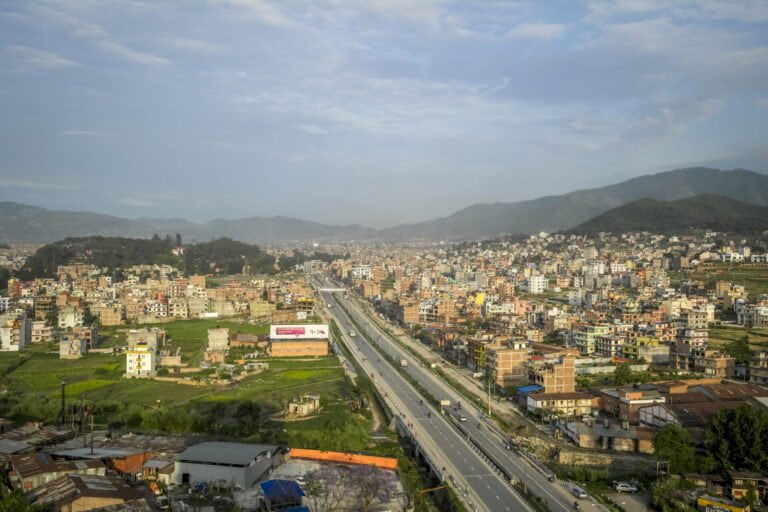 Aerial view of a bustling city with dense building clusters, a clear highway cutting through the middle, and surrounding green hills under a blue sky.