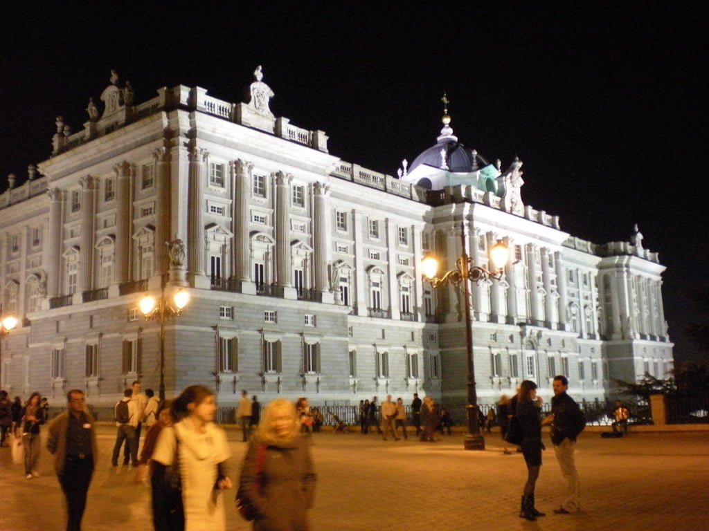 Night view of the illuminated Royal Palace of Madrid with people in the foreground.