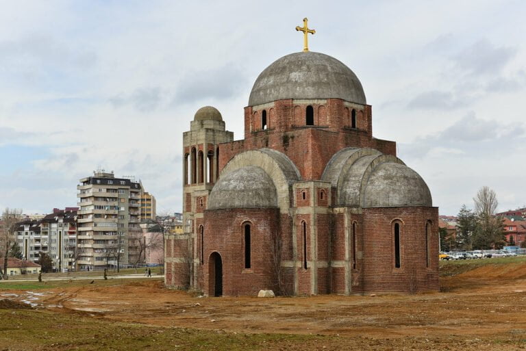 A partially constructed red-brick Orthodox church with a golden cross atop its main dome, surrounded by an undeveloped landscape with residential buildings in the background under a cloudy sky.